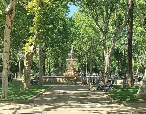 Vista de la fuente central rodeada de árboles y senderos en un parque lleno de visitantes.
