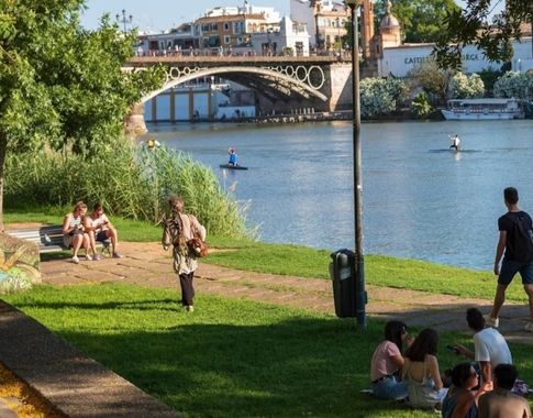 Personas disfrutando junto al río Guadalquivir en Sevilla con un puente histórico al fondo.