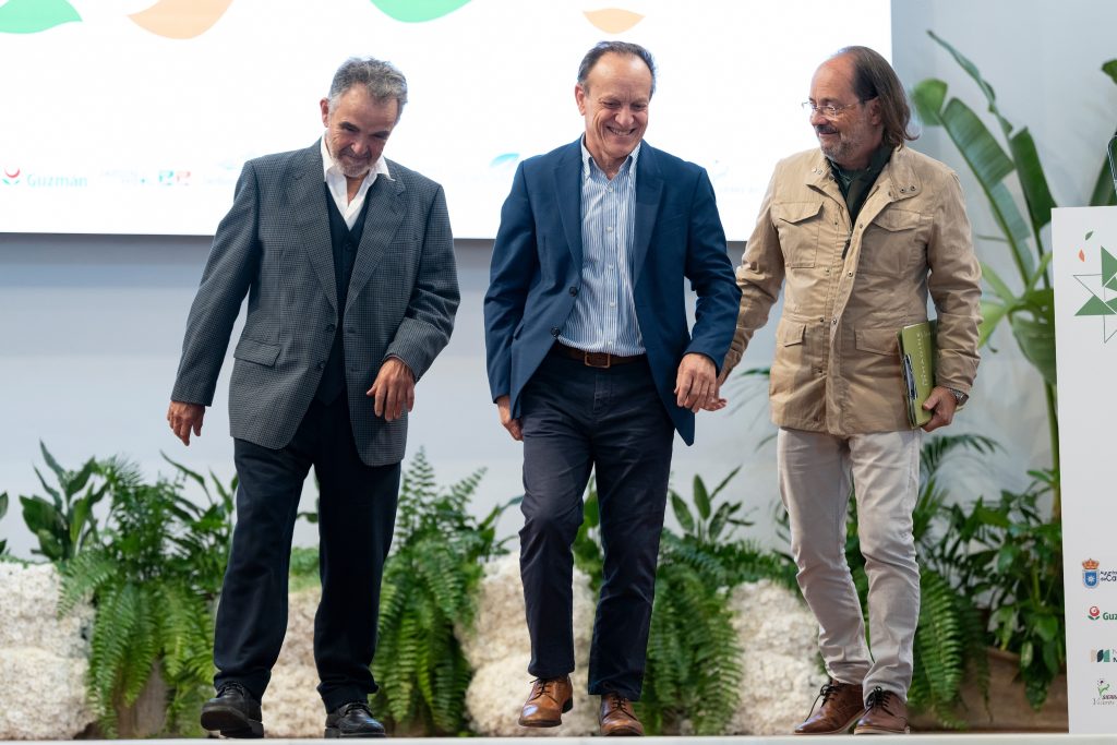Tres hombres caminando juntos en un escenario durante un evento empresarial con fondo de plantas decorativas y pantalla de presentación.