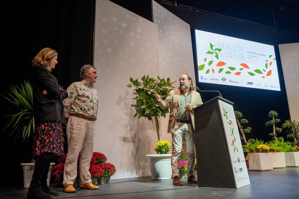 Tres personas en un escenario durante el evento de entrega de premios Andalucía en Flor, rodeados de plantas y flores.